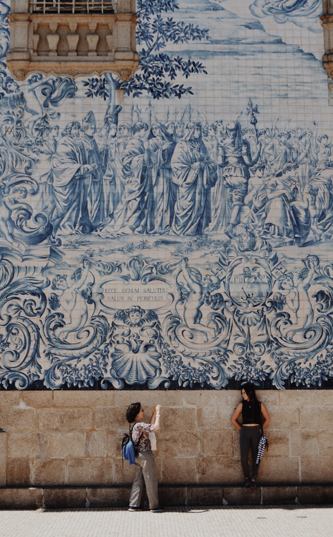 Foto de duas mulheres posando em uma parede com uma arte. Uma está tirando a foto da outra.