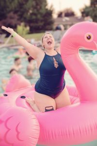 Uma mulher plus size na piscina, em cima de uma boia grande de flamingo. 