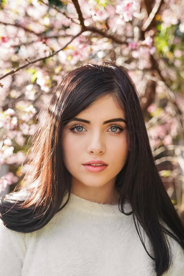 Mujer con cabello largo oscuro y ojos azules, con un árbol de flores rosas detrás de ella. Tiene un maquillaje muy colorido con azul en los ojos y labios violeta.