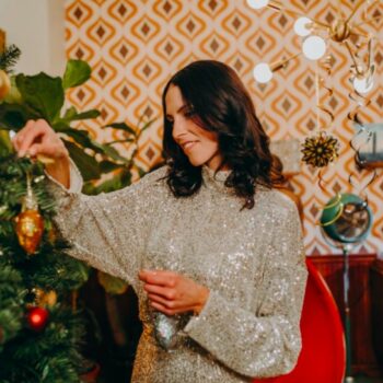 Una imagen de una mujer morena con el pelo ondulado vistiendo un cárdigan gris poniendo adornos en un árbol de Navidad.