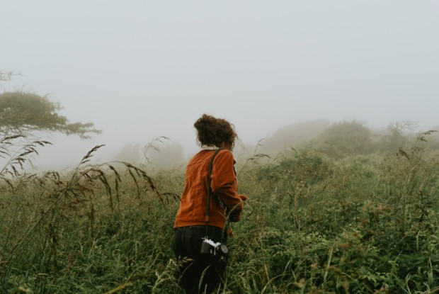 Una imagen de una joven de pelo rizado, vestida con un cárdigan naranja, caminando en medio de un campo con una cámara fotográfica.
