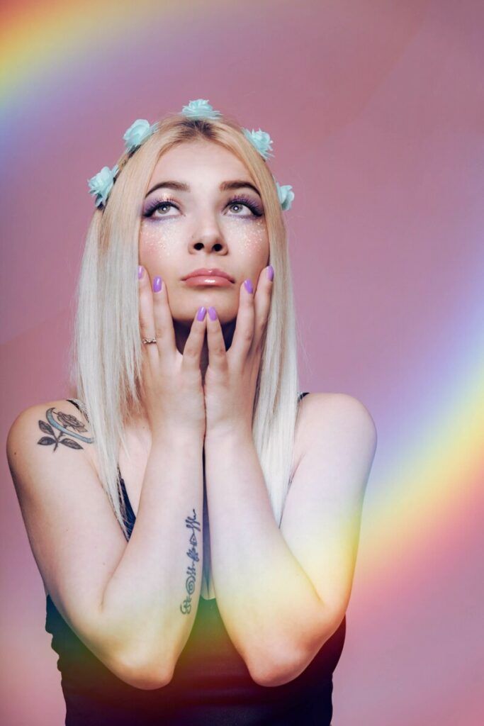 Pride Month edit of Blonde woman with blue flowers in her hair, with both hands on her face and starring up
