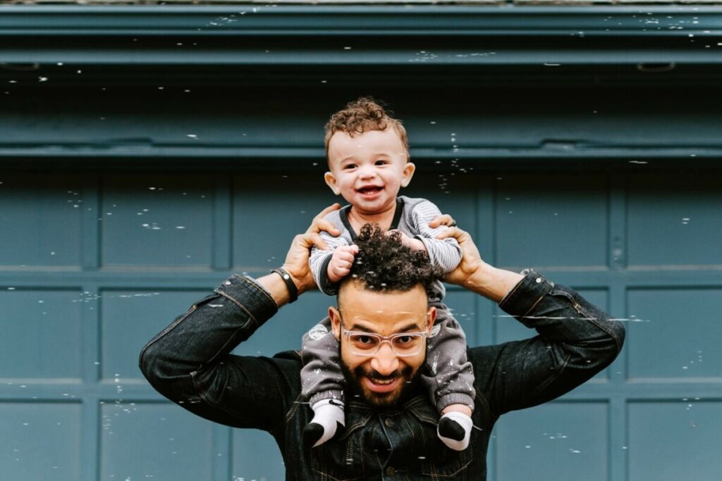 father holding his toddler son on his shoulders in front of green garage wall