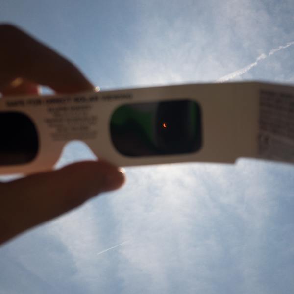 Uma mão segurando um óculos escuros diante de um eclipse solar