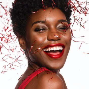 Mulher negra, em um fundo branco, comemorando e rindo, com confeites vermelho caindo sob ela