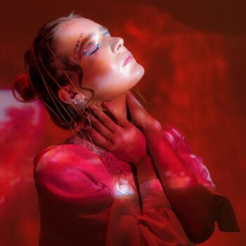 Foto embaçada de fundo vermelho e luzes vermelhas com uma mulher de olhos fechados, olhando para cima com as mãos no pescoço.