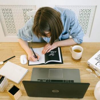 pessoa escrevendo no caderno com livros e computador na mesa